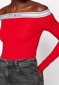 Červený body s odhalenými rameny a bílým a černým proužkovaným výstřihem s logem "TOMMY JEANS". Kombinováno s šedými džínami s vysokým pasem.