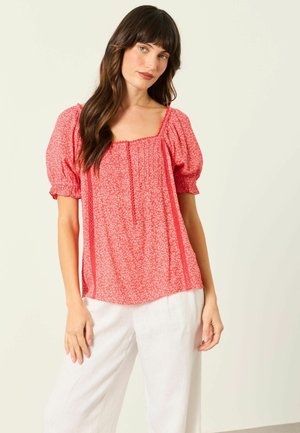 Kobieta w czerwonej, wzorzystej bluzce z bufkami i kwadratowym dekoltem oraz białych luźnych spodniach, stojąca na jednolitym jasnym tle.