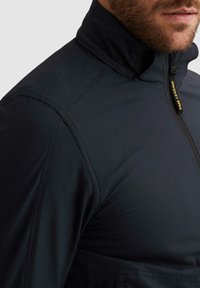 Mann trägt eine dunkelblaue Jacke mit Reißverschluss, das PME Legend-Logo ist am Reißverschlusszug in der Nähe des Kragens sichtbar.