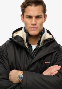 Parka negra impermeable con forro de felpa color crema, cierre de cremallera y a presión, y parche con el logo. Modelo lleva puesto un reloj con correa de cuero marrón.