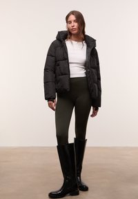 Schwarze Daunenjacke mit Kapuze, weißes Langarmoberteil, olivgrüne Leggings und knielange schwarze Stiefel. Glatte, glänzende Texturen sind vorhanden.