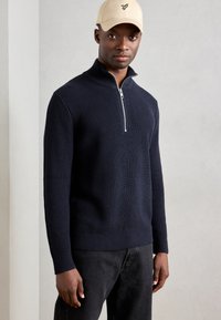 Marinefarvet strikket pullover med høj krave og halv lynlås. Ribbet struktur og manchetter, parret med mørke jeans. Beige kasket med logo.