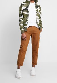 Maskáčová mikina na zip v zelené a bílé barvě, doplněná hnědými kalhotami a bílými teniskami. Mikina má žebrované manžety a vysoký límec.