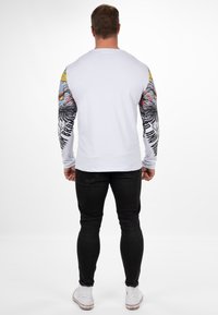 Langärmliges weißes Hemd mit bunten grafischen Ärmeln, die ein Wolfsmotiv zeigen, kombiniert mit schwarzen Jeans und weißen Sneakern.