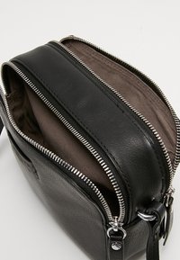 Svart läder crossbody-väska med två zippered-fack, silverdetaljer och en texturerad yta. Insidan är av en slät, neutral tyg.