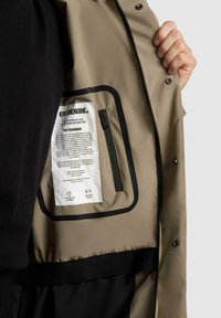 Interno di giacca beige con tasca con zip nera e etichetta di cura. Texture liscia, accenti neri a contrasto e dettagli di chiusura sicuri.