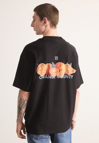 Juodas medvilninis marškinėlis su dideliu oranžiniu piešiniu ir "ORANGE COUNTY" tekstu ant nugaros. Trumpomis rankovėmis, laisvas kirpimas.