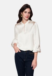 White blouse with embroidered floral design along shoulders, featuring a keyhole neckline and billowy sleeves. Paired with dark jeans.