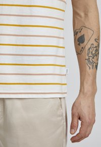Short-sleeve striped t-shirt in white, with yellow and light pink horizontal stripes. Features a side label and is paired with beige pants.