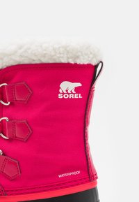 La bota impermeable fucsia cuenta con un cuello de piel sintética blanca, suela de goma negra y herrajes metálicos con un logo de oso polar y la marca "SOREL".