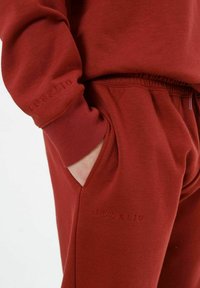 Bordeauxfarbene Sweatshirtmaterial mit einer weichen Textur, ausgestattet mit gerippten Bündchen und einem dekorativen, aufgenähten Logo am Ärmel. Sichtbare Taschen.