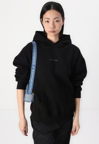 Sweatshirt negru cu glugă și buzunar frontal, prezentând logo-ul "PEGADOR". Purtați-l cu o geantă de umăr din denim cu un curea cu model.