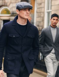 Dunkelblaue Knopfjacke mit zwei vorderen Taschen und Rippstruktur, kombiniert mit einem dunklen Hemd und einer Mütze, im Hintergrund ein in Grau gekleideter Mann.