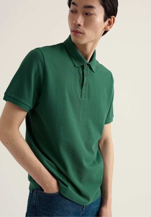 Giovane uomo con polo verde a maniche corte e jeans blu, in piedi con una mano in tasca, guarda verso destra davanti a uno sfondo semplice.