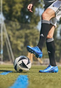 Blaue Fußballschuhe aus synthetischem Material, schwarze Kniestrümpfe und ein schwarz-weißer gemusterter Fußball auf Gras mit blauen Linien.