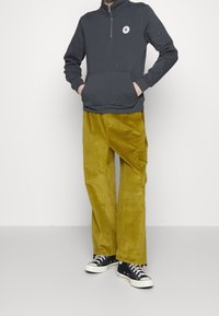 Sudadera gris oscura con cremallera y bolsillo frontal, combinada con pantalones anchos de pana amarillos. Zapatillas negras completan el atuendo.