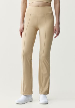 Kvinde iført beige lige bukser og hvide slip-on sneakers, stående mod en ensartet lys baggrund.