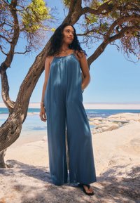 Tuta blu, ampia e comoda, in tessuto strutturato, con spalline sottili, pantaloni larghi e silhouette rilassata. Indossata con infradito nere.