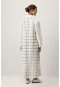 Longue robe à carreaux crème avec des lignes bleues, à coupe droite et col montant. Portée sur une chemise blanche, assortie à des chaussures blanches.