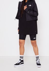 Czarna kurtka przeciwwiatrowa nałożona na czarny, oversize'owy t-shirt, zestawiona z czarnymi szortami rowerowymi oraz czarno-białymi trampkami w paski skarpetami.