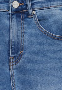 Jeans de mezclilla en un color azul medio, diseño clásico de cinco bolsillos, costuras claras y un botón de metal con detalle de marca.