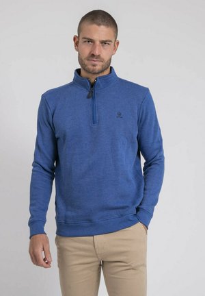 Homme aux cheveux courts portant un pull à fermeture éclair bleue et un pantalon beige, debout devant un fond clair uni.