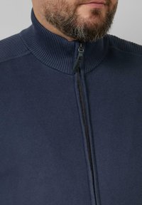 Marineblauwe trui met ritssluiting en geribde kraag, met een gladde textuur en een zwarte rits. Close-up van de halslijn.