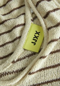 Beige stickad tyg med bruna horisontella ränder. Etiketten är ljusgul med svart text. Texturen verkar mjuk och något löst stickad.