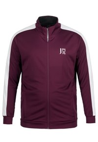 Chaqueta con cremallera en color marrón oscuro, con franjas laterales blancas, cuello alto y logo en el lado izquierdo del pecho. Fabricada con un tejido liso y elástico. Tiene dos bolsillos.