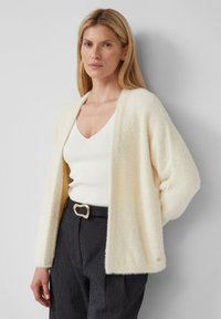 Gestructureerde crèmekleurige cardigan met open voorkant en verlaagde schouders, gedragen over een witte V-hals top. Bevat een gouden accentdetail aan de zoom.