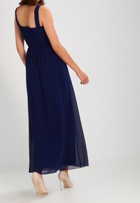 Woman wearing a sleeveless navy blue floor-length dress and nude high-heeled shoes, standing against a plain light background.