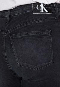 Jeans de mezclilla negra con un ajuste ceñido, que cuentan con un parche de logo en la cintura y dos bolsillos traseros con sutiles detalles de costura.