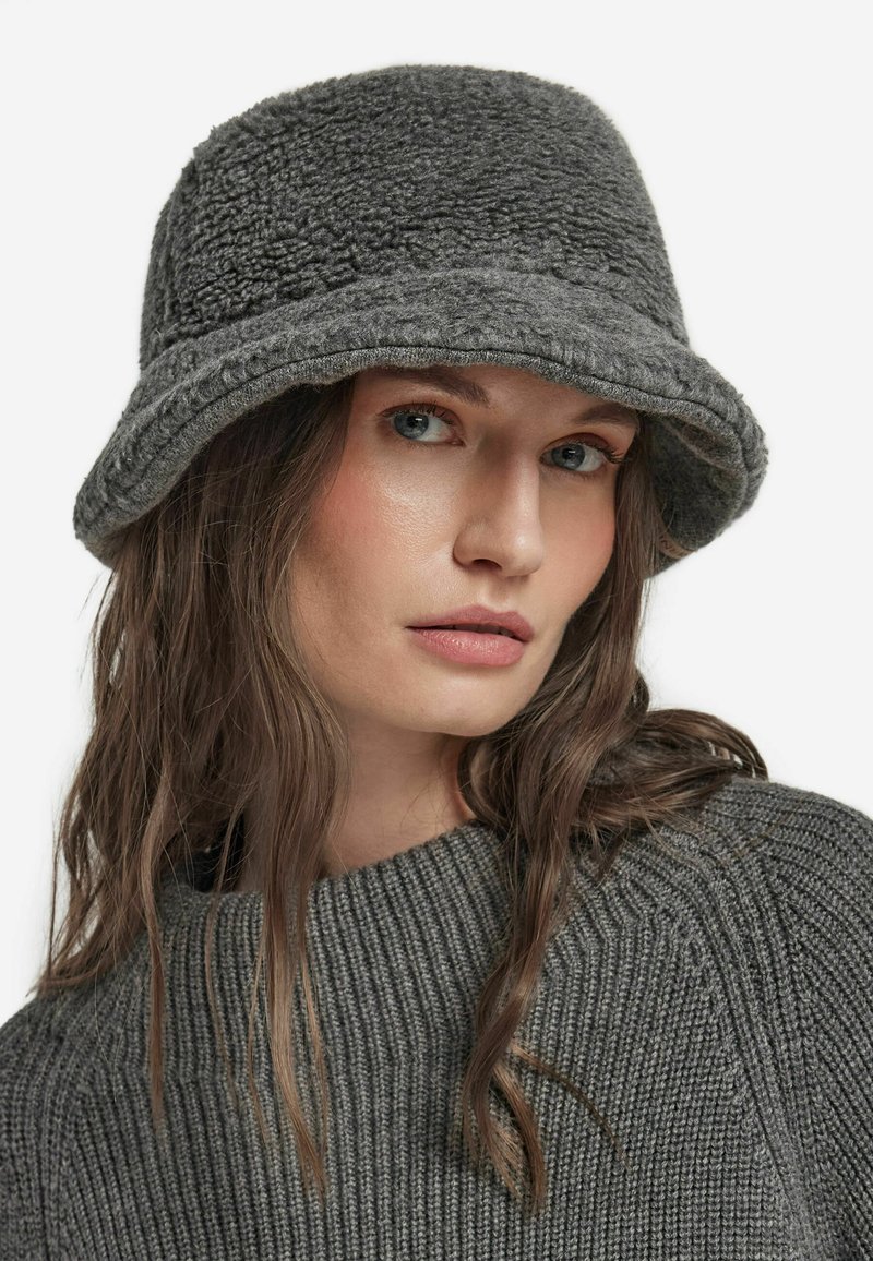 Szara wiadrowa czapka wykonana z miękkiego, teksturowanego materiału, z szerokim rondem i przeszytym brzegiem. Noszona z dopasowanym, prążkowanym szarym swetrem.