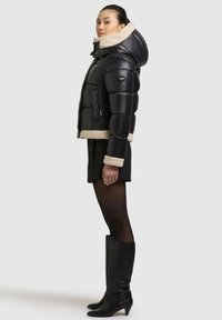 Schwarze Daunenjacke mit einem Kragen und Saum aus Kunstfell, mit einer Seitentasche und Reißverschluss. Getragen mit einem schwarzen Rock und kniehohen Stiefeln.