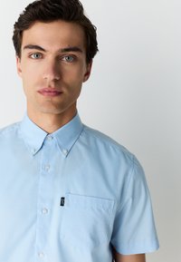 Lyseblå kortærmet skjorte med knapper og krave, med en enkelt brystlomme og karakteristisk etiket, lavet af glat stof.