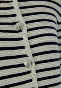 Gestreifter Strickcardigan in Creme und Marineblau. Mit runden silbernen Knöpfen mit einem detaillierten Wappen-Design. Weicher Stoff, offenes Vorderteil.