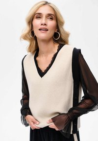 Beige gebreide vest met een zwarte bies en kantdetail bij de halslijn, gecombineerd met doorschijnende zwarte lange mouwen met ruche mouwen. Zwarte knopen aan de zijkant.