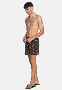Bañador marrón con un patrón multicolor de zapatillas, banda elástica en la cintura y corte relajado. Se lleva con chanclas negras, sin camiseta.