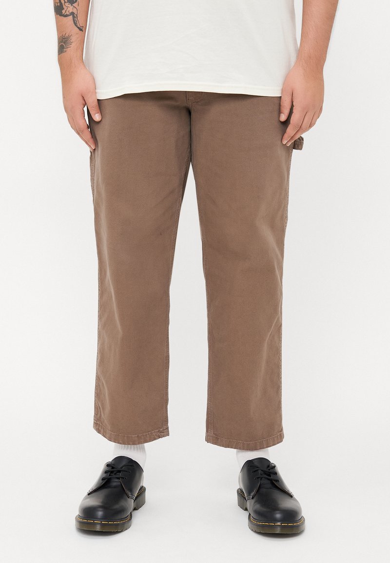 Brown cotton trousers with a relaxed fit, straight legs, and side pockets, worn with black shoes and white socks.
