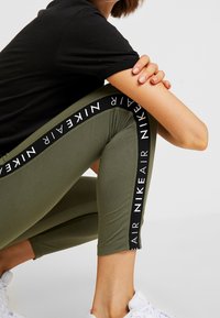 Vihreät leggingsit mustilla sivupaneeleilla, joissa on valkoista "Nike Air" -tekstiä. Valmistettu venyvästä kankaasta, lyhyet mitat ja sileä pinta.
