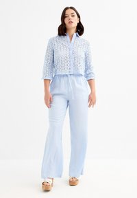 Un mono azul claro con un top de cuello, abotonado, que presenta patrones recortados y una cintura elástica, complementado por pantalones de pierna ancha.