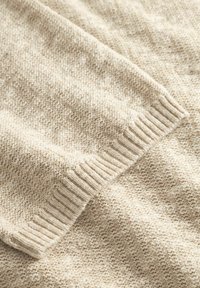 Közeli kép egy bézs színű kötött pulóver ujjáról, bordázott szegéllyel és texturált mintázattal.