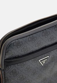 Schwarze Tasche aus synthetischem Material mit strukturiertem Logo-Muster, silbernem Reißverschluss und einem dreieckigen metallischen Markenanhänger auf der Vorderseite.