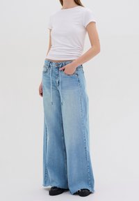 Pantalones vaqueros de pierna ancha de color azul claro con cintura alta, con cordones, dos bolsillos frontales y dobladillo deshilachado. Combinados con una camiseta blanca ajustada.