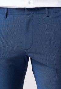 Schmal geschnittene blaue Hose aus strukturiertem Stoff, mit flachem Vorderteil und seitlichen Taschen mit glatter Oberfläche.
