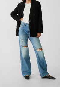 Person wearing a white top, black blazer, ripped wide-leg blue jeans, and black pointed-toe shoes, standing against a plain background.