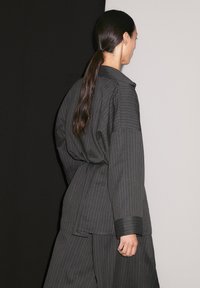 A woman with long, dark hair faces away, wearing a black-and-white striped outfit, against contrasting black and white backgrounds.