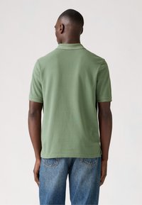 Short-sleeve polo shirt in olive green cotton, featuring a flat collar and subtle stitching details, paired with blue denim jeans.