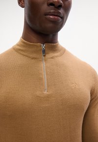 Maglione lavorato a maglia color beige con colletto alto e chiusura a mezza zip in argento. Presenta un piccolo logo ricamato sul lato sinistro. Texture morbida.