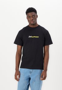 Black cotton t-shirt with short sleeves, featuring a small yellow and white logo on the front. Styled with light blue jeans.
